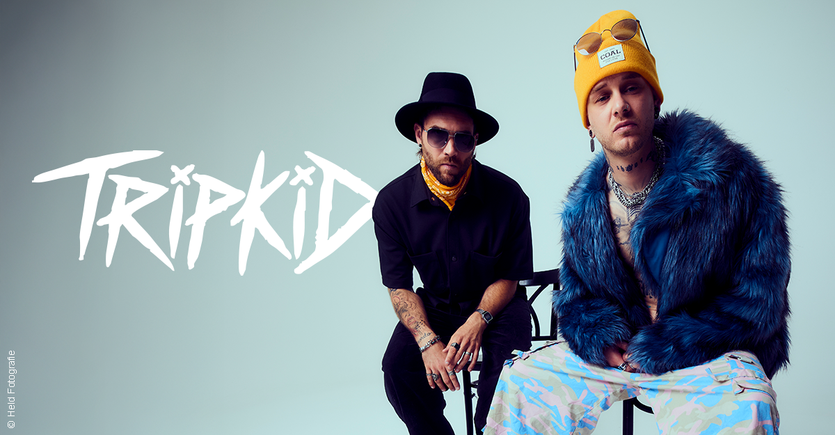 Zwei Männer in auffälliger Kleidung posieren vor einem hellen Hintergrund. Einer trägt einen Hut und Sonnenbrille, der andere eine gelbe Mütze und blaue Jacke.