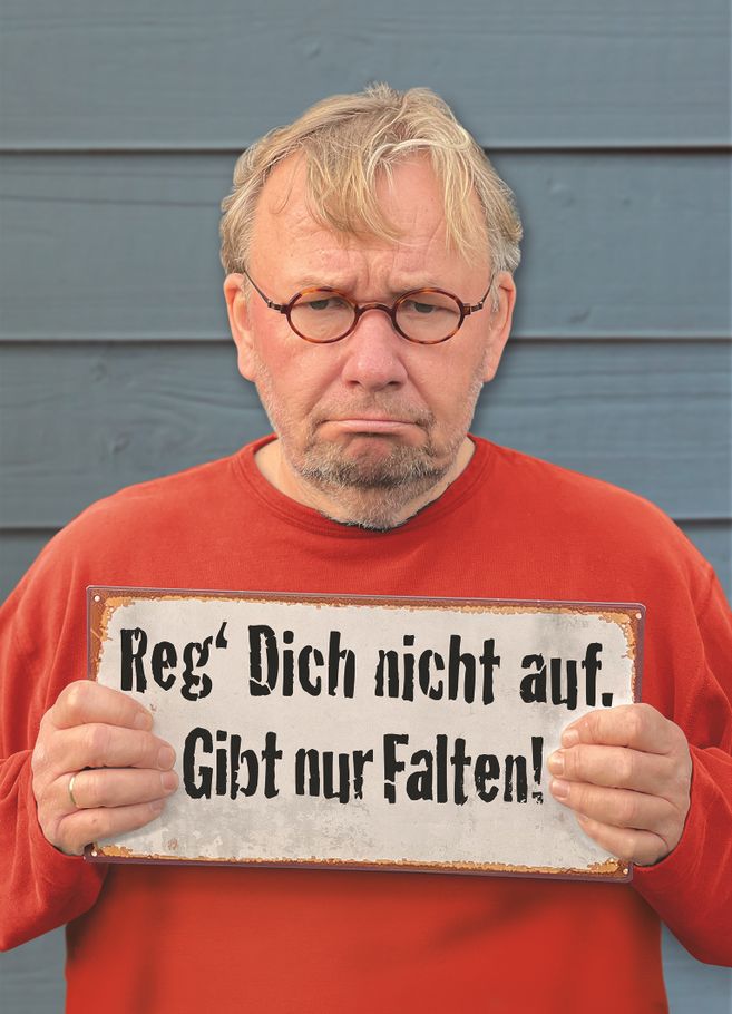 Ein Mann mit Brille und rotem Pullover hält ein Schild mit der Aufschrift: 'Reg' Dich nicht auf. Gibt nur Falten!' vor einer blauen Wand.
