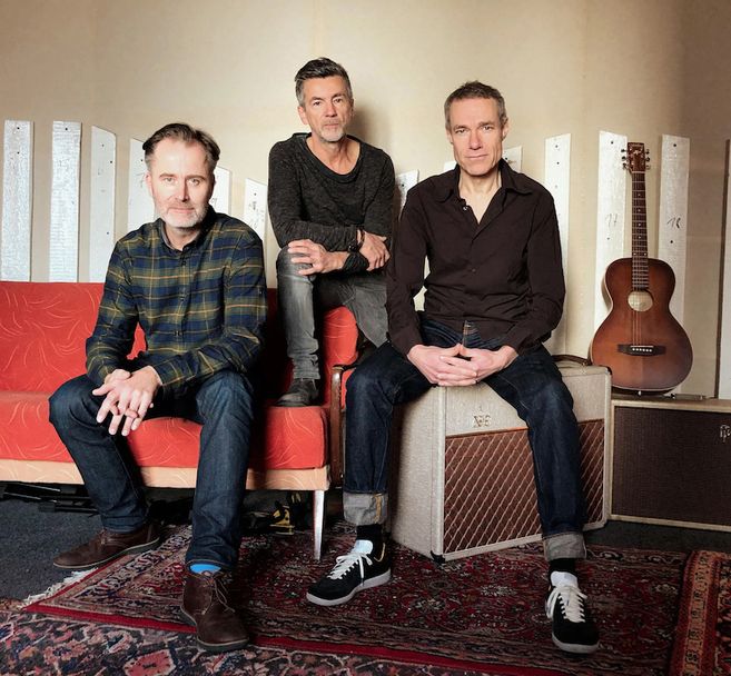 Drei Männer sitzen in einem Raum mit einer Gitarre im Hintergrund.