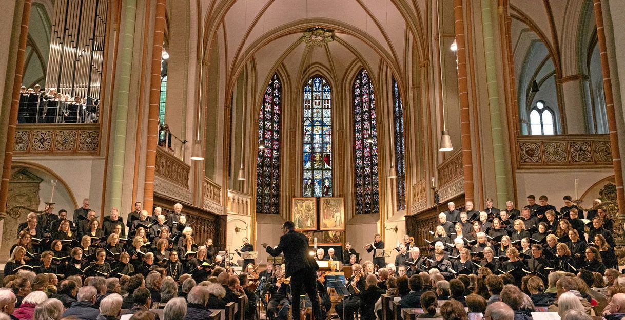 Ein Chor und Orchester führen in einer Kirche ein Konzert auf. Der Dirigent steht vor dem Ensemble, während das Publikum in den Bänken sitzt.