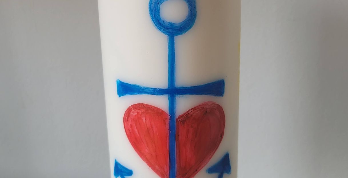 Eine brennende weiße Kerze mit einem blauen Anker und einem roten Herz darauf.