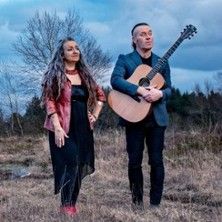Zwei Personen stehen auf einem Feld, eine Frau und ein Mann mit Gitarre.