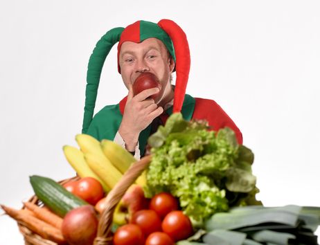 Ein Mann in einem bunten Narrenkostüm isst einen Apfel hinter einem Korb voller Obst und Gemüse.