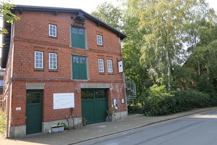 Backsteingebäude mit grünen Türen und Fenstern, umgeben von Bäumen.