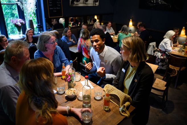 Menschen sitzen in kleinen Gruppen an Tischen und tauschen sich in einem Café über verschiedene Themen aus. Im Vordergrund ist eine Gesprächsrunde zu sehen.