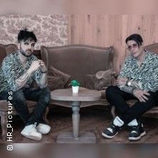 Zwei Personen sitzen auf Sesseln vor einem Holztisch in einem Raum mit Holzwänden.