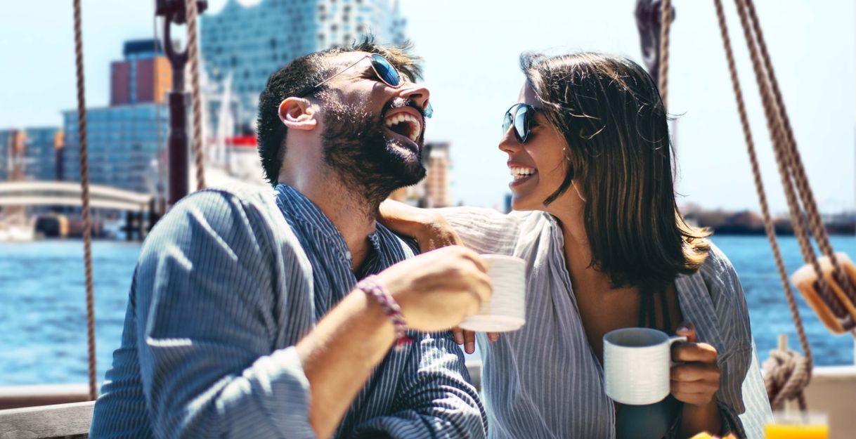 Ein Pärchen sitzt an Deck eines Segelschiffes beim Brunch. Im Hintergrund ist die Elbphilharmonie zu sehen.