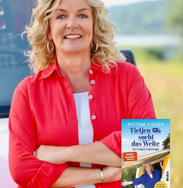 Eine Frau in roter Bluse steht lächelnd vor einem Camper. Ein Buchcover mit dem Titel 'Tietjen sucht das Weite' ist eingeblendet.