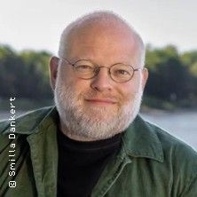 Porträt eines Mannes mit Brille und Bart vor einer natürlichen Kulisse.