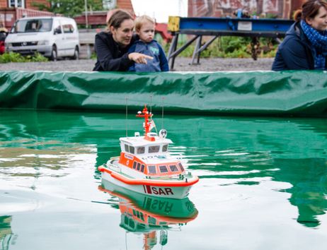 Ein Modellschiff schwimmt in einem Wasserbecken, während eine Frau und ein Kind es beobachten.