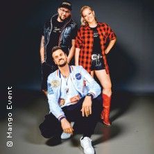 Drei Personen posieren vor einem neutralen Hintergrund. Eine Person sitzt vorne, zwei stehen dahinter. Alle tragen lässige Kleidung.