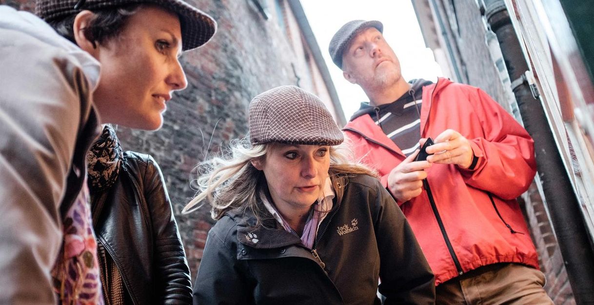 Drei Personen in Detektivkleidung in einer engen Gasse, die etwas untersuchen.