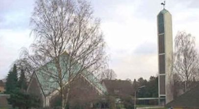 Kirche mit modernem Turm und spitzem Dach, umgeben von Bäumen.