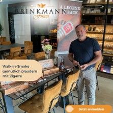 Ein Mann steht in einer Zigarrenlounge neben einem Tisch mit Zigarren und einem Werbebanner.