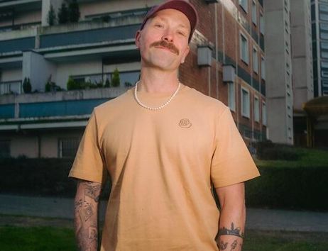 Ein Mann mit Schnurrbart und Tattoos steht vor einem Wohngebäude.