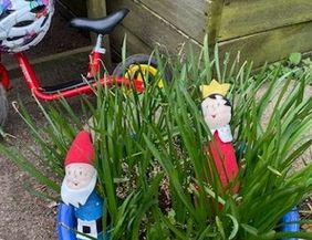 Ein rotes Kinderfahrrad steht neben einem Pflanzentopf mit Gras, in dem zwei Holzfiguren, ein Zwerg und ein König, stecken.