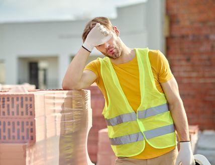Ein Bauarbeiter in einer gelben Warnweste wischt sich den Schweiß von der Stirn. Er steht neben einem Stapel Ziegelsteine auf einer Baustelle.