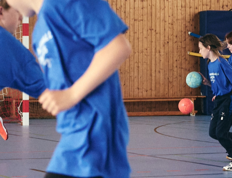 Kinder in blauen T-Shirts, die einen Handball prellend durch eine Sporthalle laufen
