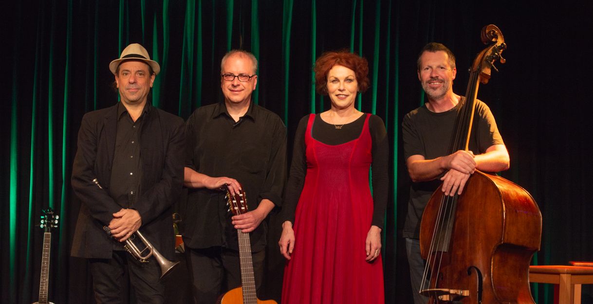 Vier Musiker stehen auf einer Bühne vor einem grünen Vorhang. Sie halten Instrumente wie Trompete, Gitarre und Kontrabass.