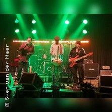 Drei Musiker auf einer Bühne mit grüner Beleuchtung, einer spielt Saxophon, ein anderer Gitarre und der dritte Bass.