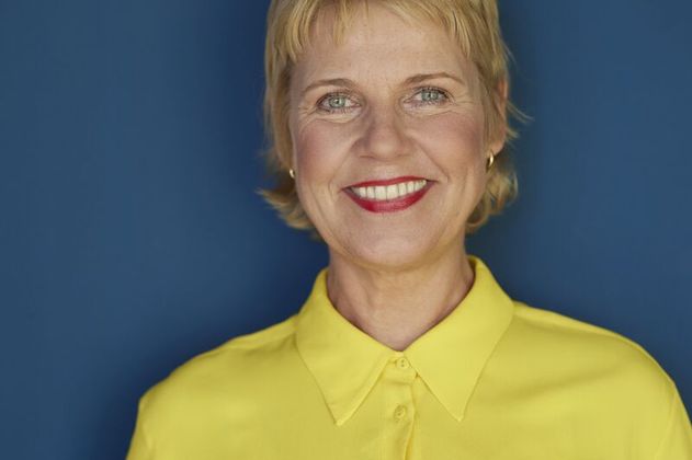 Frau in gelber Bluse vor blauem Hintergrund lächelt in die Kamera.