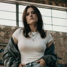 Eine Frau in einem weißen T-Shirt und einer offenen Jacke steht vor einer Backsteinwand.