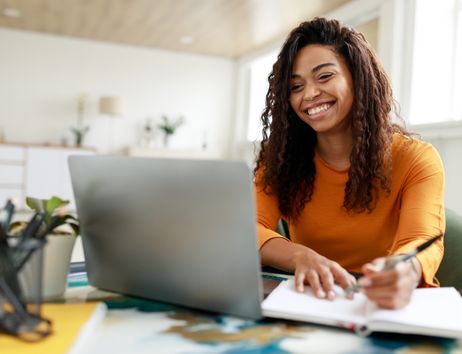 Junge Frau sitzt lächelnd an einem Tisch vor einem Laptop, macht sich Notizen in einem Notizbuch. Der Raum ist hell und freundlich, mit großen Fenstern im Hintergrund