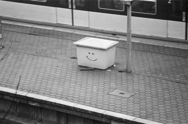 Auf einem Bahnsteig steht ein kasten, auf den ein Smiley gesprüht ist. Im Hintergrund fährt eine alte S-Bahn. Das Bild ist aus den 90ern und in schwarz-weiss.