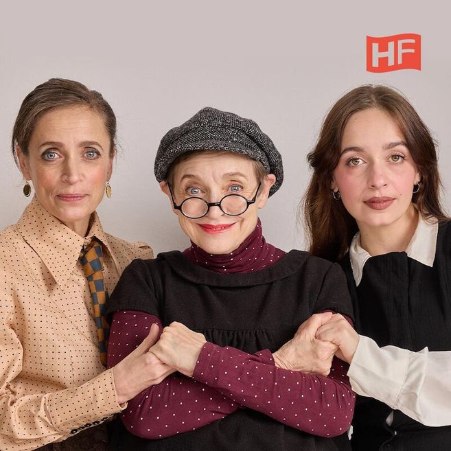 Drei Frauen posieren zusammen, die mittlere trägt eine Brille und Mütze, die anderen zwei haben lange Haare. Im Hintergrund ist ein HF-Logo zu sehen.
