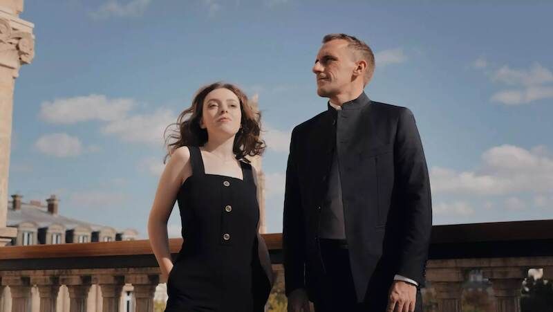 Ein Mann und eine Frau stehen auf einem Balkon mit einem historischen Gebäude im Hintergrund, in Schwarz-Weiß aufgenommen.