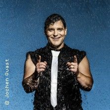 Ein Mann in schwarzer Kleidung und weißer Krawatte steht vor einem blauen Hintergrund und lächelt, während Wasser auf ihn fällt.