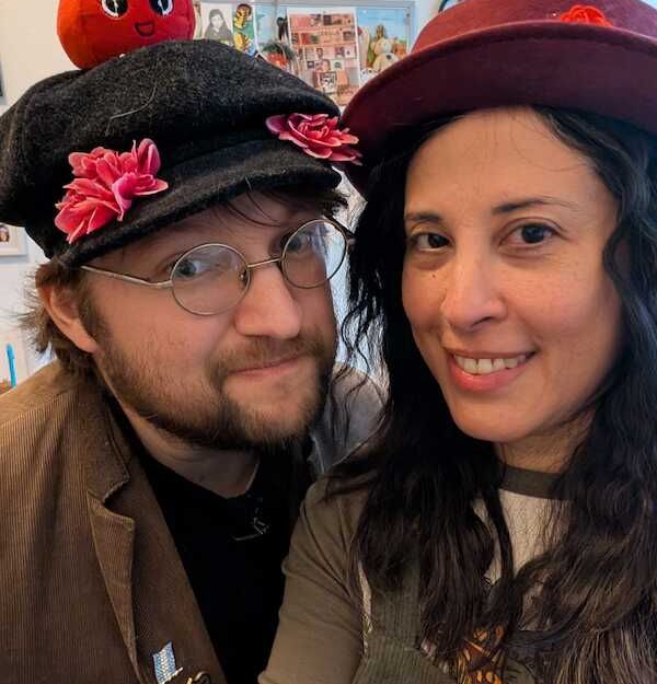 Zwei Personen mit Hüten, einer mit einem Apfel auf dem Hut, lächeln in die Kamera.