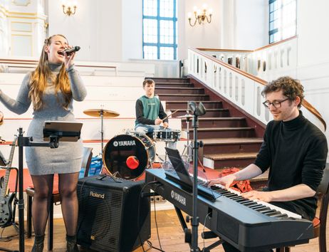 Eine Jugendband probt in einer Kirche. Eine Sängerin singt, während ein Schlagzeuger und ein Keyboardspieler musizieren.