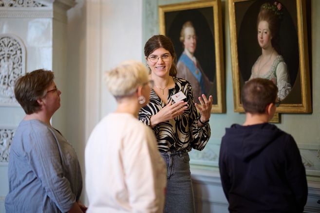 Eine Frau führt eine Gruppe durch ein Schloss und erklärt Gemälde an der Wand.