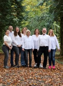 Eine Gruppe von acht Personen steht auf einem mit Herbstlaub bedeckten Waldweg. Sie tragen weiße Hemden und lächeln in die Kamera.