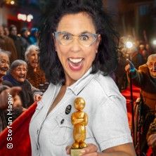 Eine Frau mit Brille hält eine goldene Trophäe, umgeben von jubelnden Menschen auf einem roten Teppich.