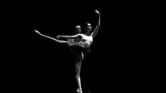 Ein Ballettpaar tanzt auf einer dunklen Bühne, der Mann hebt die Frau in einer eleganten Pose.