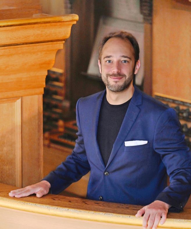 Ein Mann in einem blauen Anzug steht an einer Holzbalustrade und lächelt in die Kamera.