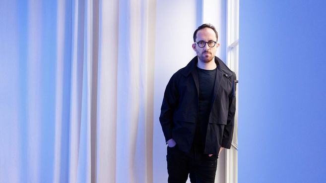 Eine Person in schwarzer Kleidung steht vor einem blauen Hintergrund.