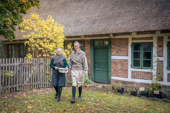 Zwei Frauen in historischer Kleidung vor einem reetgedeckten Fachwerkhaus mit grünem Türrahmen und herbstlichem Garten.
