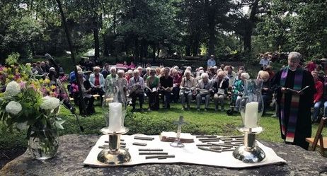 Eine Gruppe von Menschen sitzt im Freien vor einem Altar mit Kerzen und Blumen, während ein Geistlicher eine Zeremonie abhält.