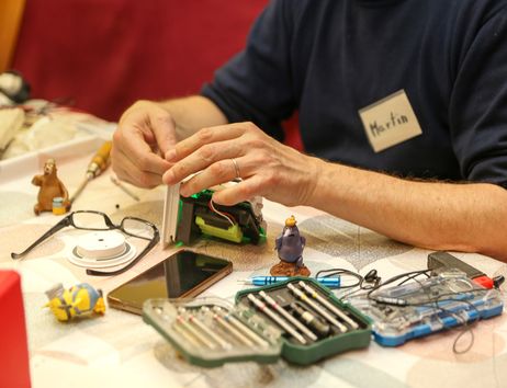 Person repariert ein elektronisches Gerät an einem Tisch mit Werkzeugen und kleinen Figuren.