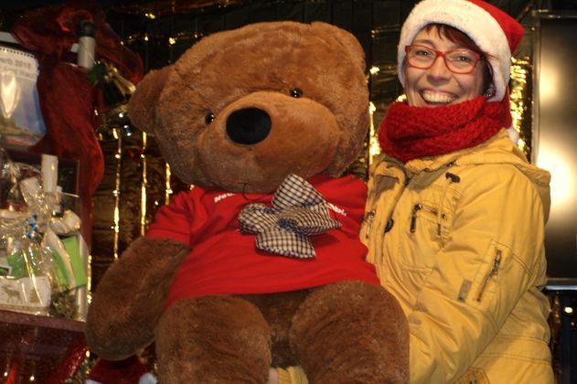 Eine Person mit Weihnachtsmütze hält einen großen Plüschbären auf einem Weihnachtsmarkt.
