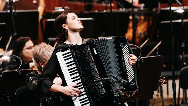Eine Frau spielt Akkordeon auf einer Bühne mit einem Orchester im Hintergrund.