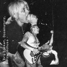 Schwarz-Weiß-Foto von Musikern auf der Bühne, einer singt leidenschaftlich ins Mikrofon, ein anderer spielt Gitarre.