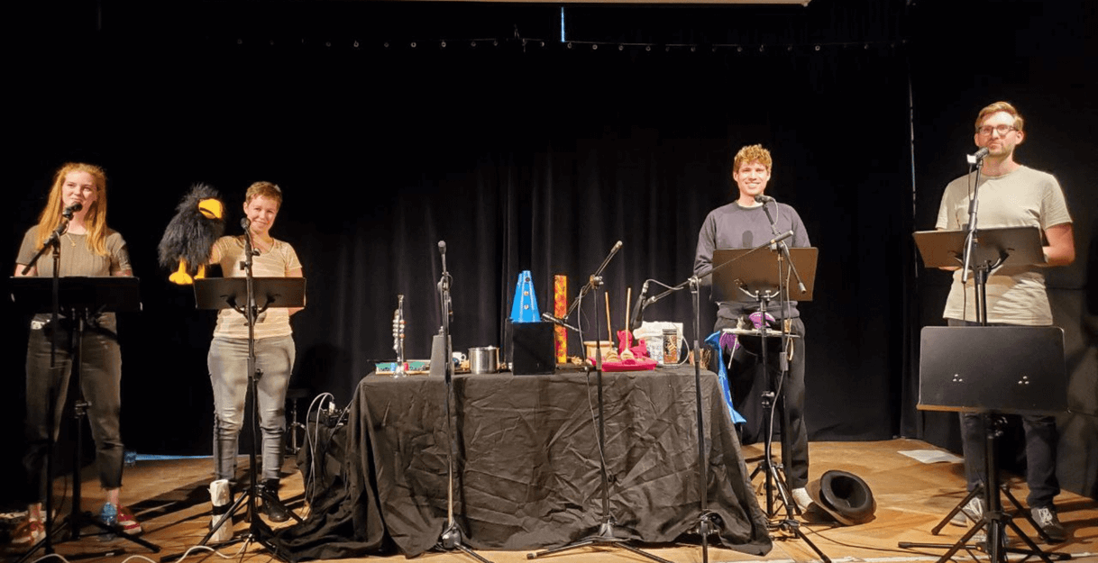 Vier Personen stehen auf einer Bühne mit Mikrofonen und Requisiten für ein Live-Hörspiel.
