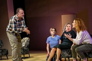 Vier Personen in einem Theaterstück, eine steht und gestikuliert, drei sitzen und hören zu.