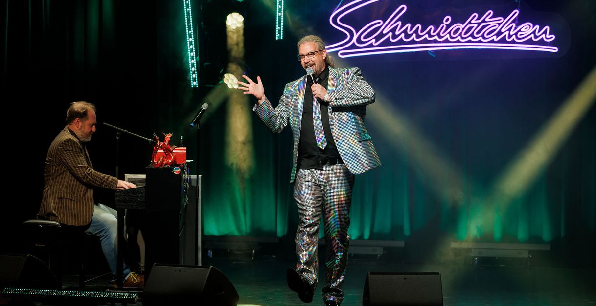 Ein Mann in einem glitzernden Anzug singt auf einer Bühne, während ein anderer Mann Klavier spielt. Im Hintergrund leuchtet ein Schild mit der Aufschrift 'Schmidtchen'.