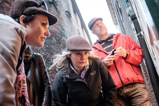 Drei Personen in Detektivkleidung in einer engen Gasse, die etwas untersuchen.