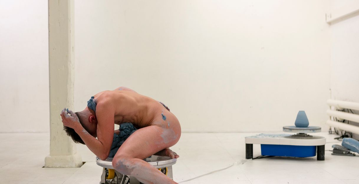 Excerpt from Simone Aughterlony's performance "Hand Job." A naked person sits with their head buried in their hands, covered in a clay-like substance, on a stool and can only be seen from the side.
